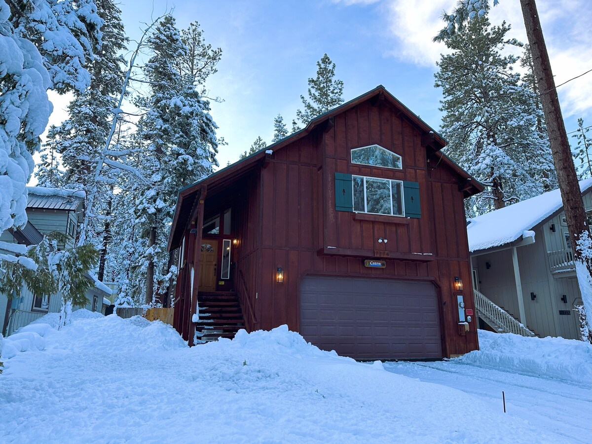 A two-story cabin in a wooded setting reflects a rustic mountain style. The exterior features wooden siding with green shutters and a large garage door, surrounded by a blanket of snow. Tall pine trees stand nearby, enhancing the peaceful ambiance.