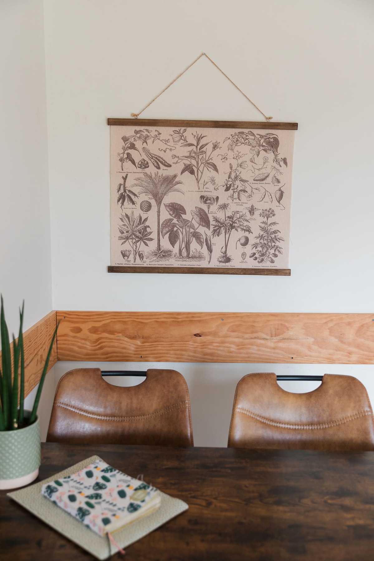 A wall-mounted botanical print is displayed above a wooden dining table. Two brown leather chairs are positioned at the table, complemented by a green potted plant nearby. A decorative notebook with a floral cover rests on the table's surface.