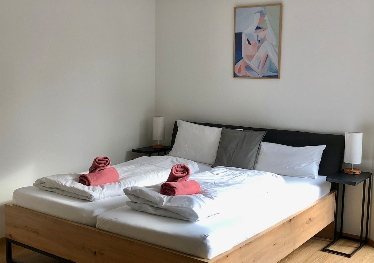 A spacious sleeping area features a large, neatly arranged bed with white linens. Two rolled towels in red are placed on top of the bedding. Nightstands with soft lamps are situated on either side, and a colorful artwork is displayed on the neutral-toned wall.