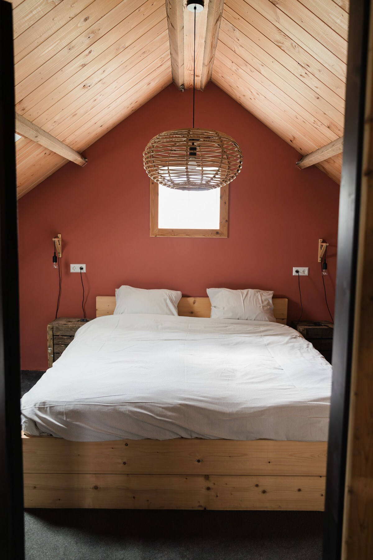 A cozy bedroom features a spacious bed with white bedding, set against a warm, terracotta wall. Natural wood accents are present in the bed frame and surrounding nightstands. A stylish pendant light hangs above, while a window allows natural light to enter.