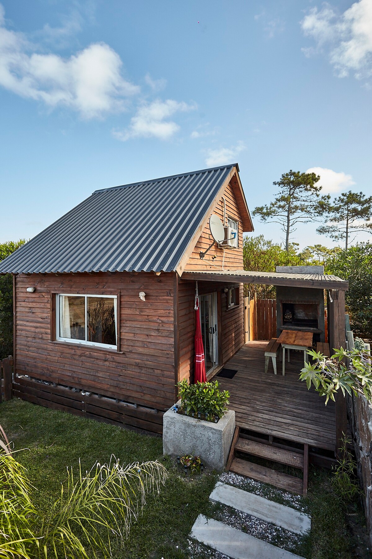A charming wooden cabin features a sloped metal roof and a front porch. A small table with chairs is situated on the porch, surrounded by greenery. Large windows allow natural light to enter, while a satellite dish is mounted on the side of the building.