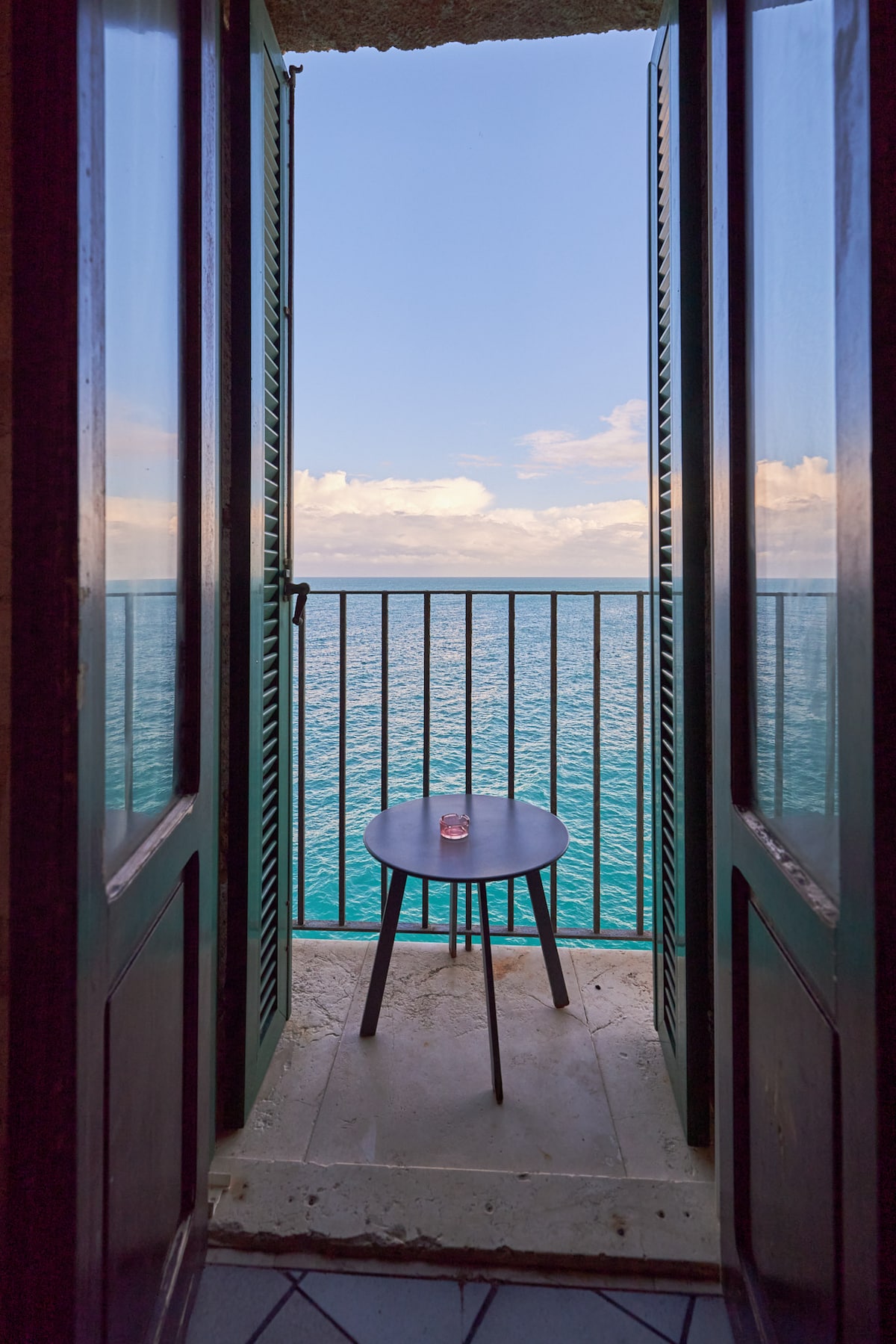 A serene balcony view is captured through open green shutters, revealing a small table with a glass placed on it. The turquoise sea extends to the horizon, meeting a bright sky decorated with soft clouds, creating a tranquil seaside atmosphere.