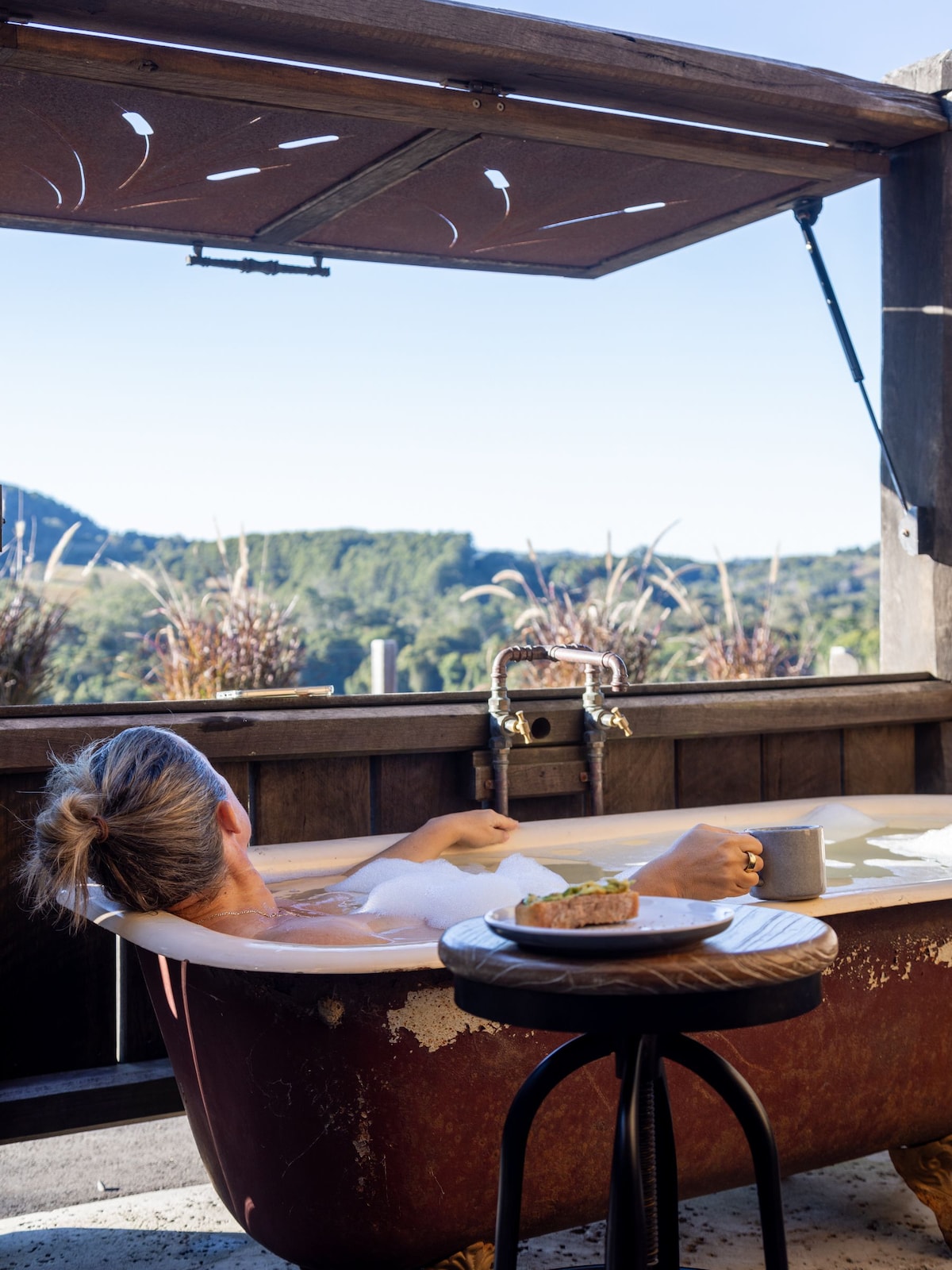 A vintage-style bathtub is positioned near an open window, offering panoramic views of the surrounding countryside. A plate of food and a cup are placed on a small wooden stool beside the bathtub, enhancing the serene setting.