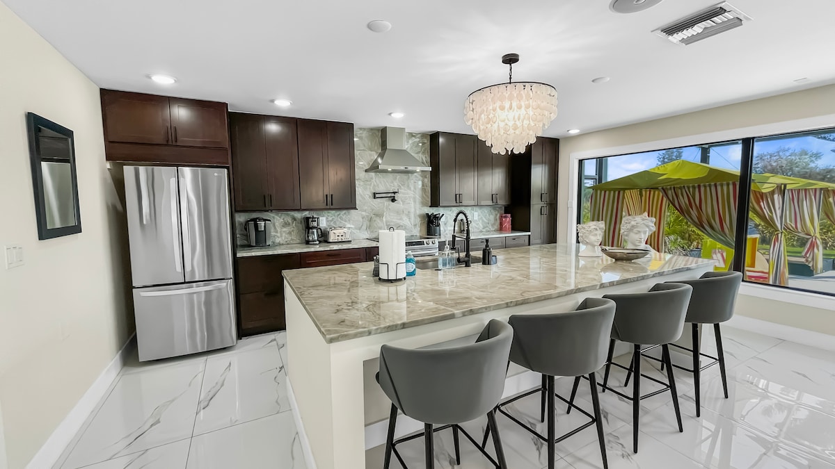 A modern kitchen features dark wood cabinetry and a large island with seating for six. Stainless steel appliances and a stylish chandelier enhance the contemporary look. A large window reveals outdoor seating with two yellow umbrellas, providing a bright, inviting view.