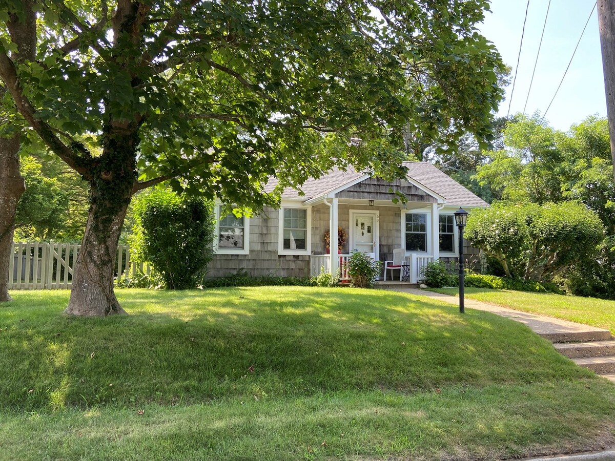 A charming cottage is surrounded by lush green grass and mature trees. The exterior features light gray shingle siding with white trim around the windows. A welcoming front porch is visible, with seating arranged for outdoor relaxation. The driveway leads up to the entrance.