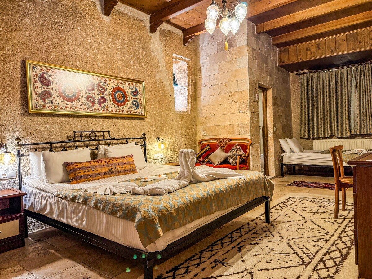 A spacious room features a king-size bed and a single bed, both adorned with soft linens. Natural stone walls create a rustic ambiance. A decorative tapestry hangs above the beds, and a wooden chair is positioned nearby. Soft lighting is provided by hanging fixtures, enhancing the warmth of the space.
