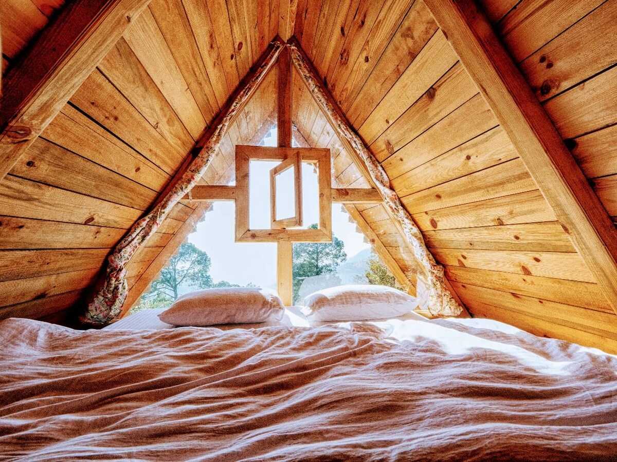 A cozy sleeping area features a spacious bed situated beneath a high, wooden sloped ceiling. Natural light filters through a unique triangular window, highlighting the textured wooden walls and inviting a serene ambiance.