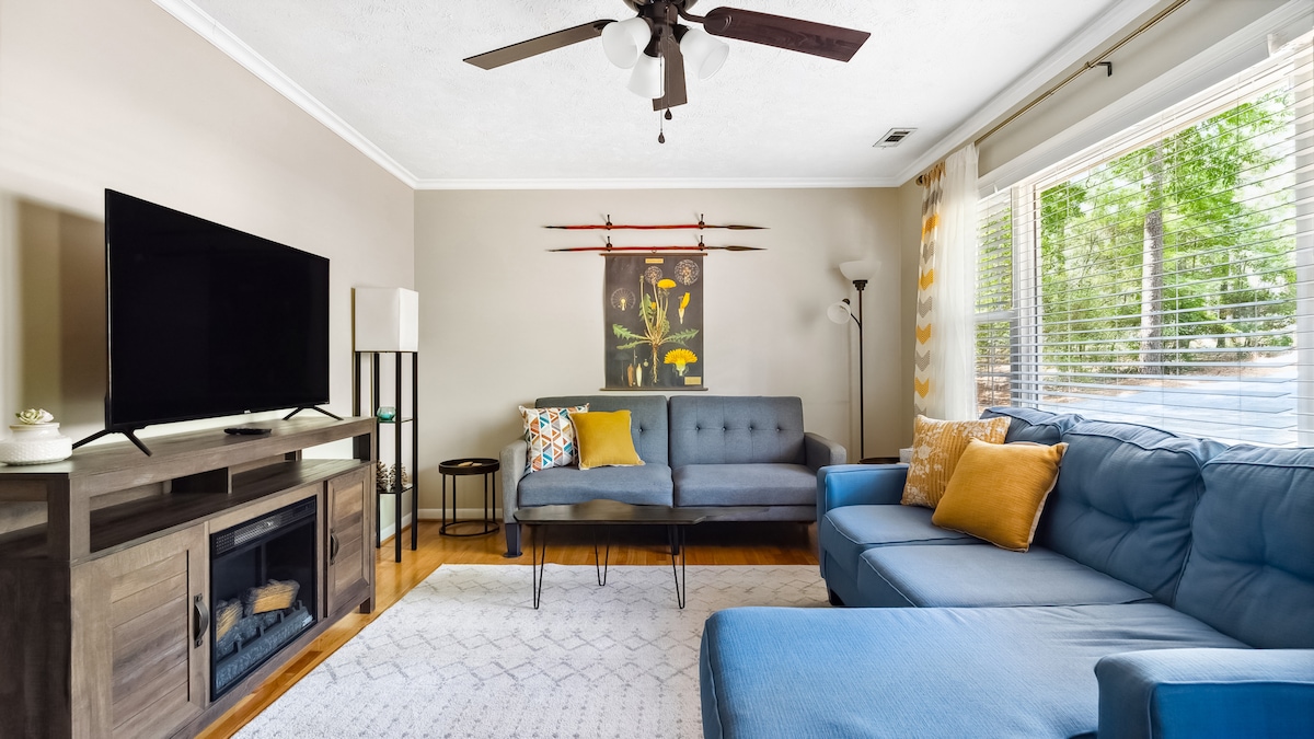 A comfortable living room features a flat-screen TV mounted on the wall, alongside a stylish electric fireplace. A blue sectional sofa and a dark gray loveseat provide seating options. Natural light enters through the window, accented by cheerful throw pillows and wall art.