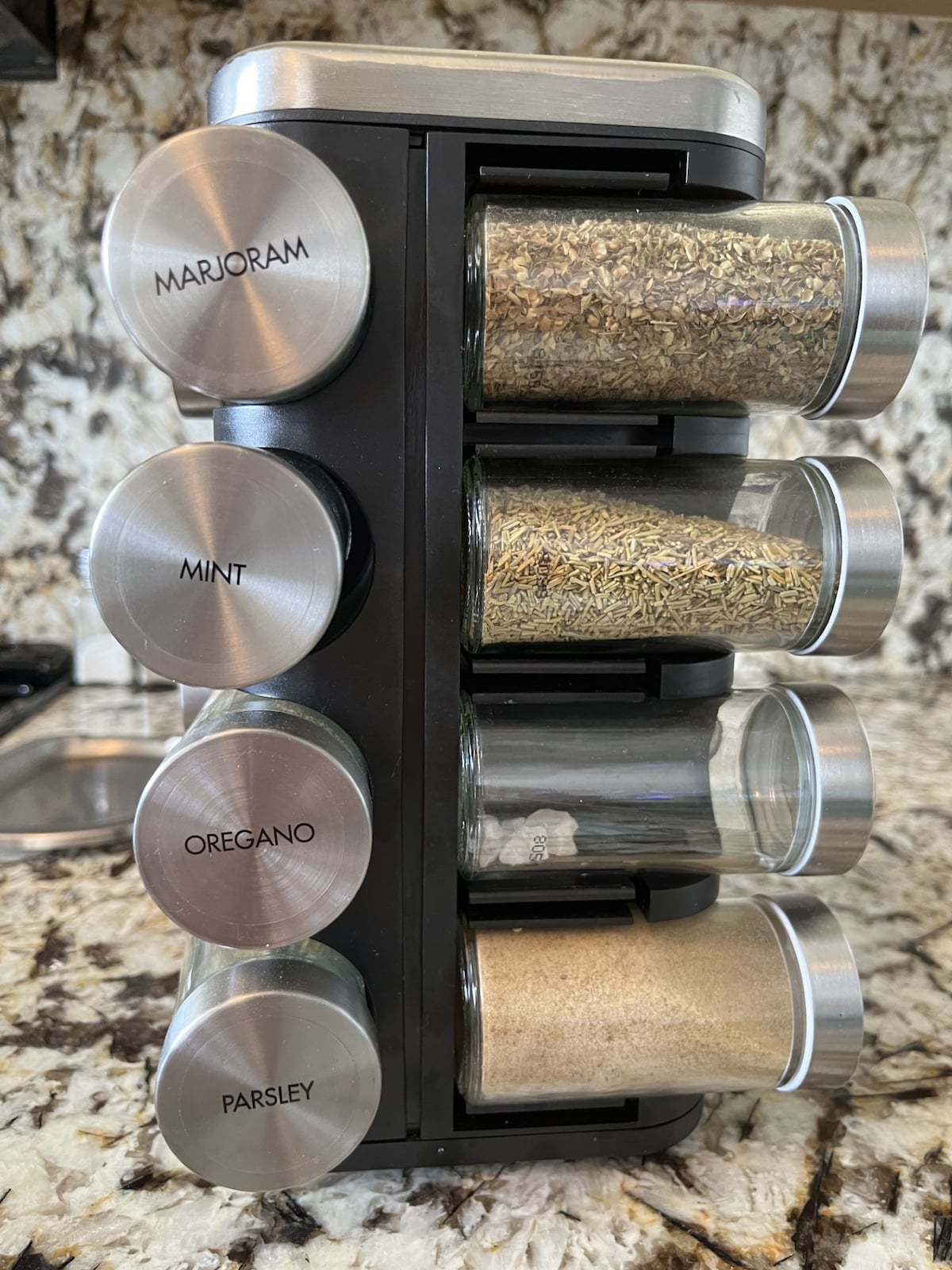 A spice rack is displayed vertically, featuring glass containers with metal lids. The labels indicate marjoram, mint, oregano, parsley, and other spices. The countertop has a textured surface, complementing the sleek design of the spice rack in a well-appointed kitchen.