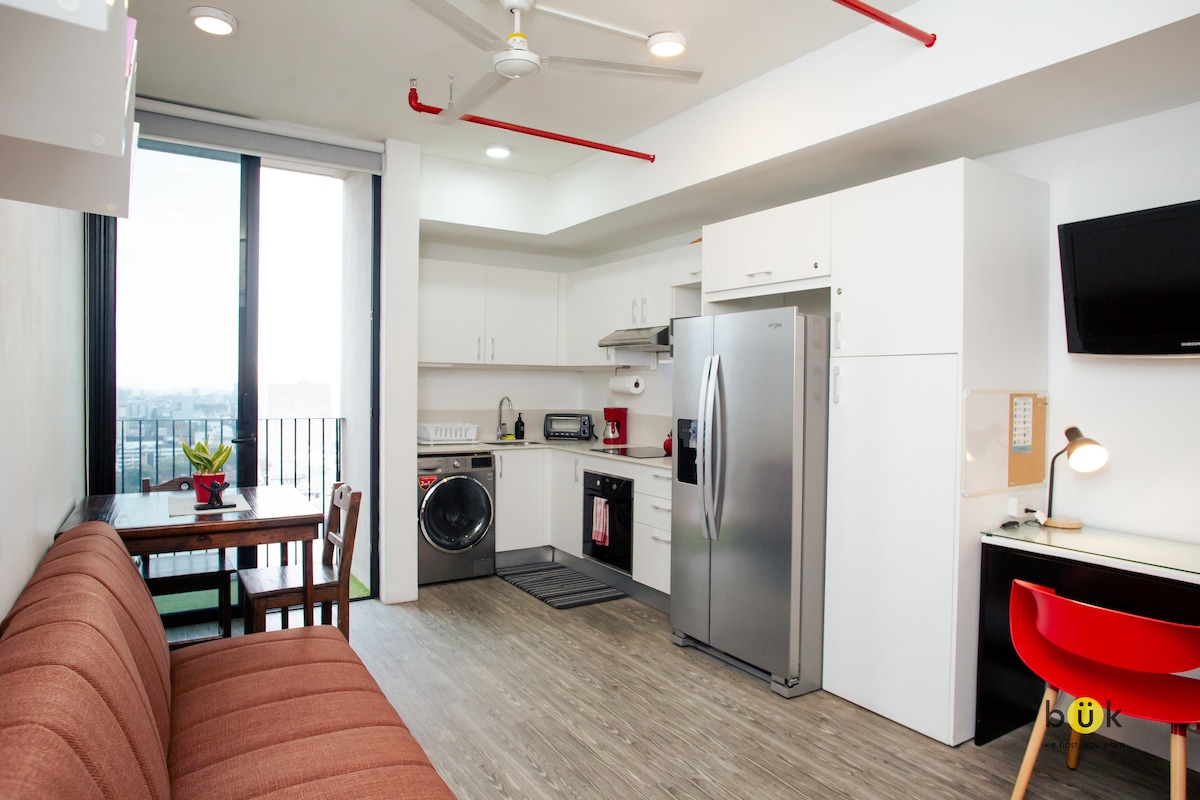A modern kitchen and living area are presented, featuring a spacious sofa, a wooden dining table, and a washer. Stainless steel appliances and white cabinetry create a clean, functional space. Large windows allow natural light to illuminate the area, with views of the surrounding city.