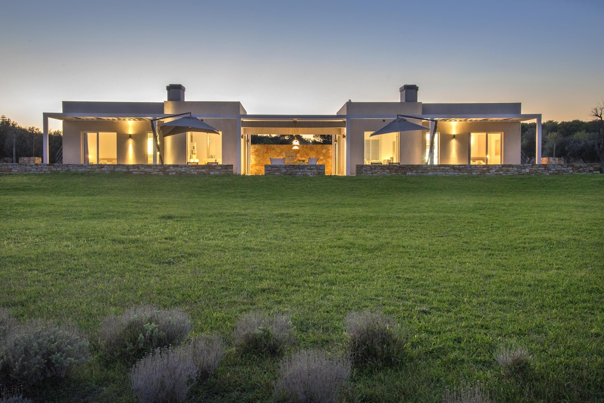 The villa is framed by a spacious green lawn, showcasing a modern architectural design with wide overhangs. Soft lighting illuminates the open patio areas on both sides, creating an inviting space for outdoor gatherings. A serene backdrop of nature enhances the tranquil setting.