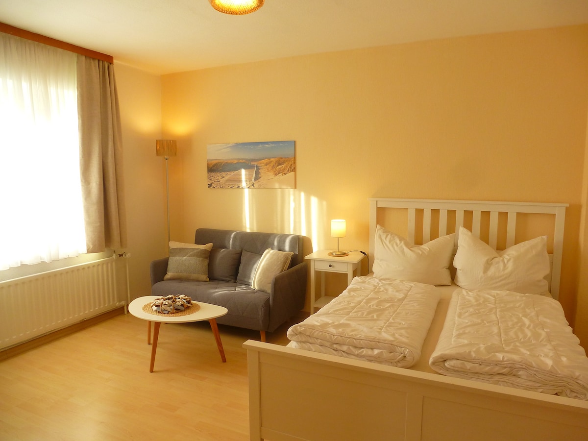 A combined living and sleeping area is featured, highlighting a spacious bed with neatly arranged bedding. A cozy sofa sits next to a modern coffee table, complemented by a soft lamp and wall art. Natural light fills the room through a large window covered with sheer curtains.