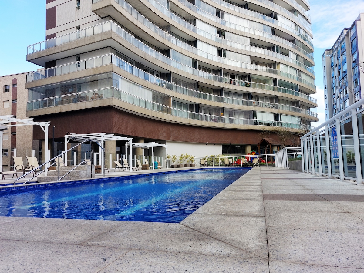 A spacious pool area is featured, surrounded by lounge chairs under shaded structures. The building's modern architecture is highlighted, with continuous glass railings providing views of the water. The area is designed for relaxation and community enjoyment.