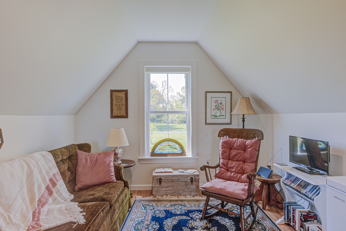 A cozy seating area featuring a plush sofa and a comfortable rocking chair. A small lamp provides soft lighting beside the large window, which frames views of the surrounding fields. A decorative rug anchors the space, enhancing the inviting atmosphere.