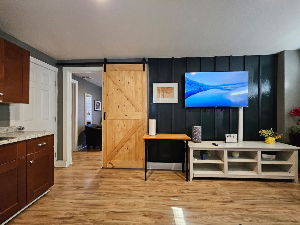 The interior features a living area with a mounted television displaying serene landscapes. A wooden barn door adds a rustic touch to the room. Modern cabinetry is visible to the left, complemented by laminate flooring that enhances the space's brightness.