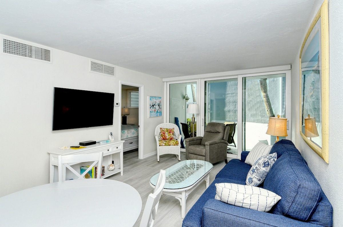 The open living area features a comfortable blue sofa and a glass coffee table, with a flat-screen TV mounted on the wall. A round dining table and chairs occupy one corner, while large glass doors reveal views of the exterior. Natural light fills the space.