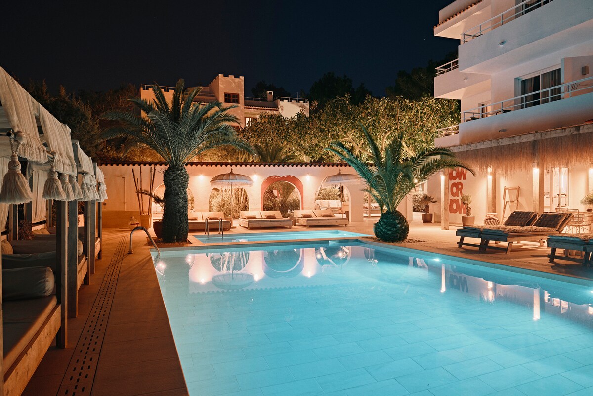 The illuminated pool area features a turquoise pool surrounded by lush palm trees and comfortable lounge furniture. Soft lighting accentuates the relaxing atmosphere of the space. The backdrop includes elegant structures with arches, contributing to the tranquil ambiance of the evening setting.