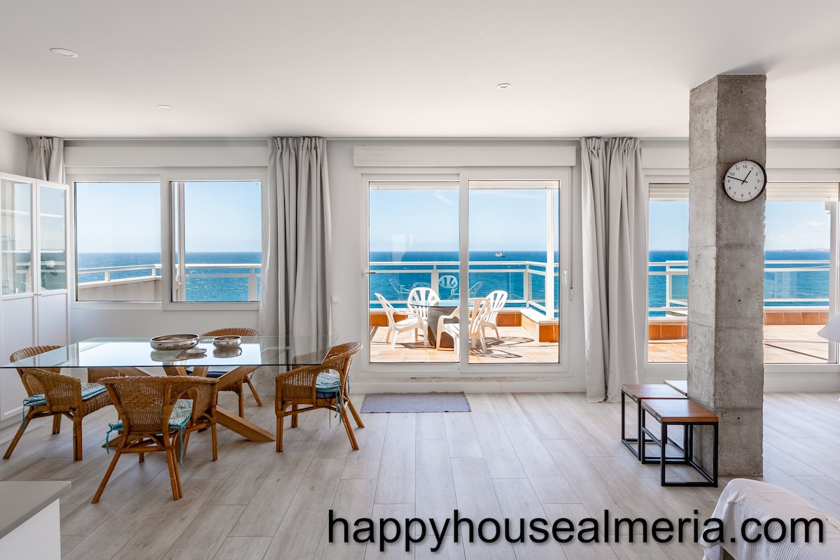 A spacious dining area features a glass table surrounded by woven chairs, with natural light flooding in from large sliding doors. A view of the terrace and the ocean beyond is visible, enhancing the open feel of the space.