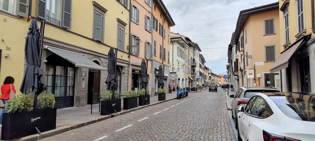 A historic street lined with charming buildings featuring colorful façades. Wide cobblestones create a rustic path, while outdoor seating areas are framed by tall planters. Parked vehicles are visible along the side, and pedestrians can be seen enjoying the environment.