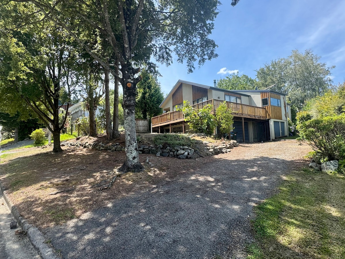 A family home is shown from the street, surrounded by green trees and manicured landscaping. The house features expansive decks on multiple sides, enhancing its connection with the outdoor environment. A gravel driveway leads up to the entrance, creating a welcoming approach.