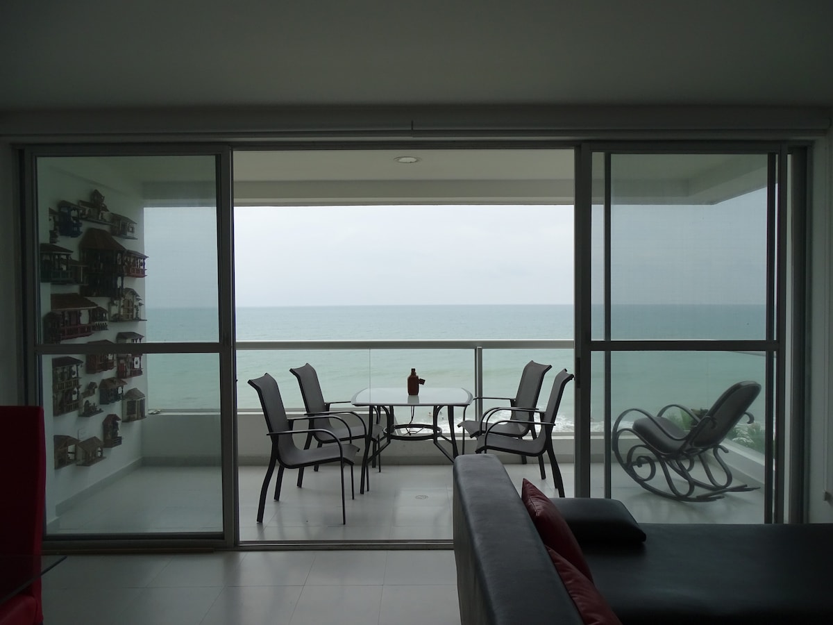 A spacious balcony features a glass table surrounded by six chairs, facing a serene ocean view. The ocean's horizon blends with the cloudy sky, enhancing the tranquil atmosphere. A rocking chair is visible in the corner, inviting relaxation while enjoying the coastal scenery.
