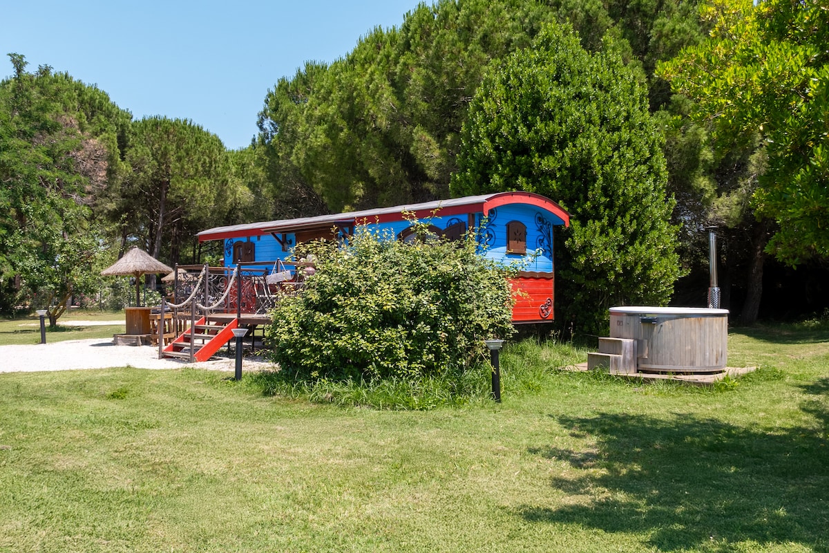A colorful gypsy caravan is set amidst lush greenery, featuring vibrant painted designs. A hot tub is positioned nearby, surrounded by well-kept grass. The path leading to the caravan is accented with stepping stones, adding to the serene outdoor environment.