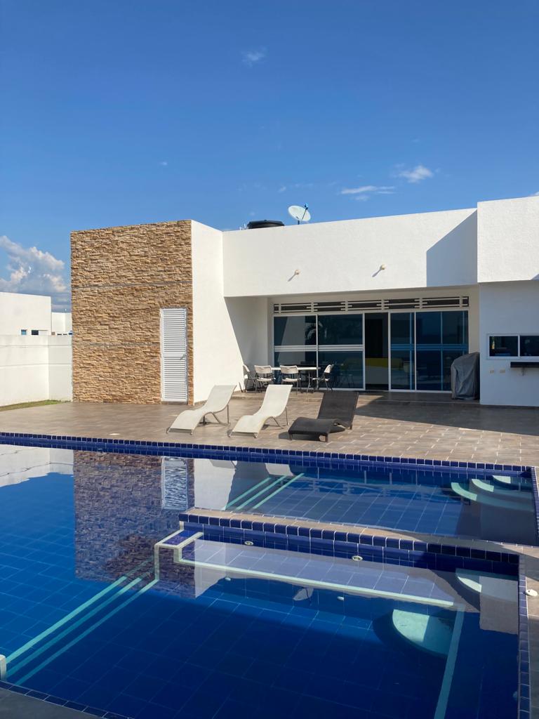 A modern home features a spacious terrace with a shimmering pool in front. Comfortable lounge chairs are positioned by the edge, and a dining area is visible in the background. The bright blue water reflects the clear sky, creating a serene ambiance.