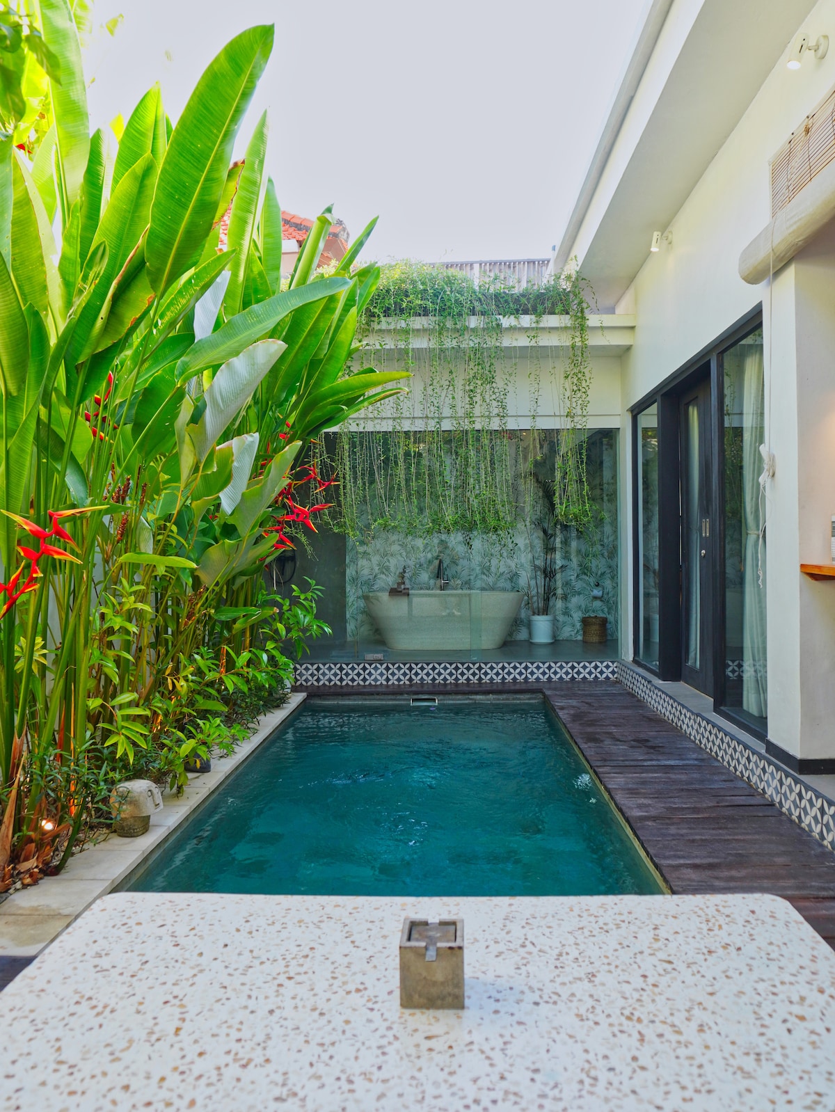 A private swimming pool is surrounded by lush tropical plants and greenery, enhancing the serene atmosphere. The modern villa features large glass doors that provide a clear view into the space, while a decorative stone surface is positioned in the foreground.