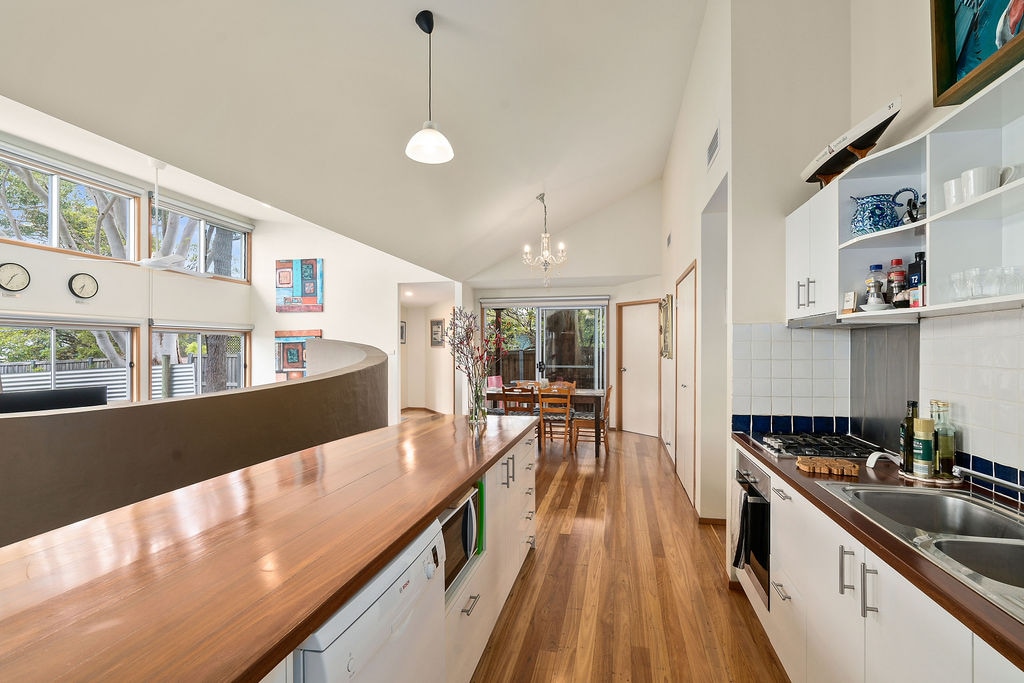 A bright kitchen area features a long wooden countertop and white cabinetry, complemented by warm wooden floors. Large windows allow natural light to fill the space. A dining table is visible in the background, with a cozy chandelier hanging above.