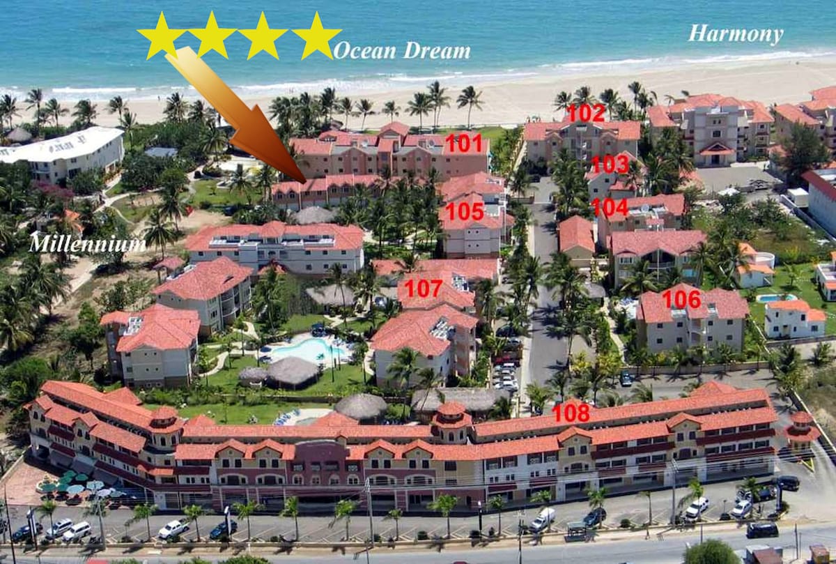 An aerial view captures a well-appointed residential complex situated close to the beach, featuring multiple buildings with terracotta roofs arranged neatly. Several swimming pools can be seen among the lush palm trees, with pathways leading to each unit. The proximity to the ocean is visually evident.