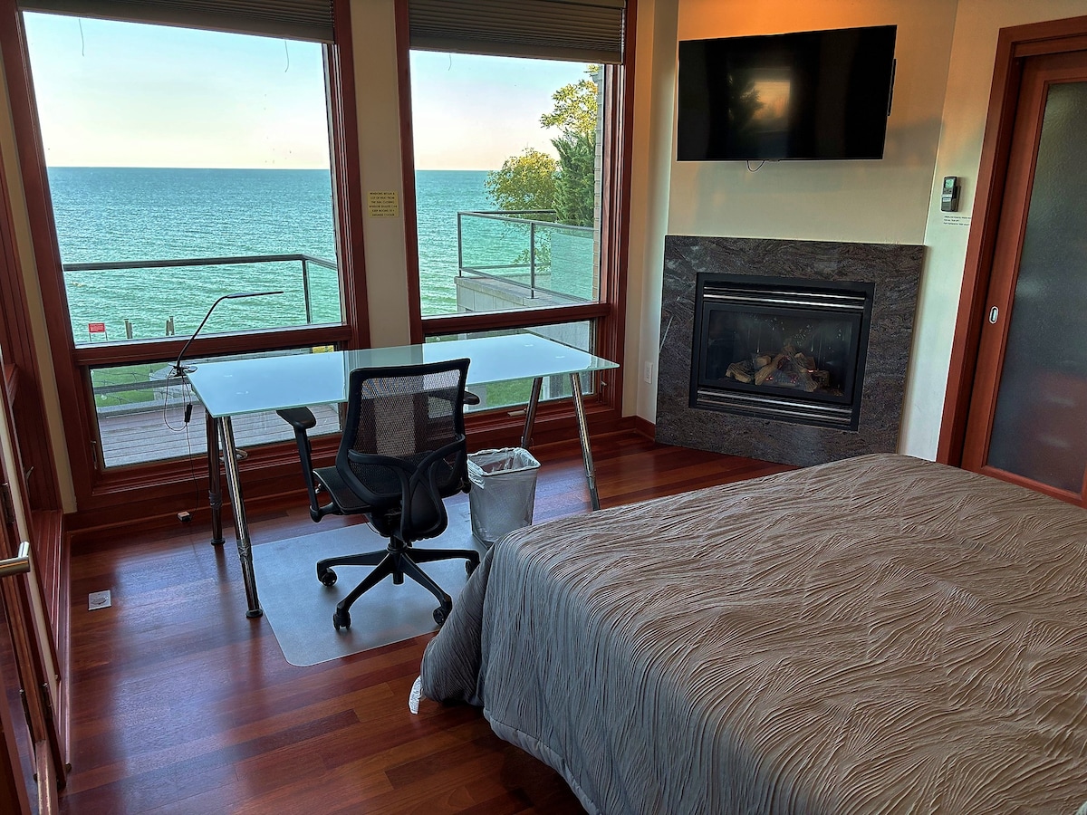 A spacious bedroom features a large window with stunning views of Lake Michigan, allowing natural light to fill the room. A comfortable bed with a textured coverled sits beside a modern desk and chair, while a wall-mounted television and elegant fireplace are also present.