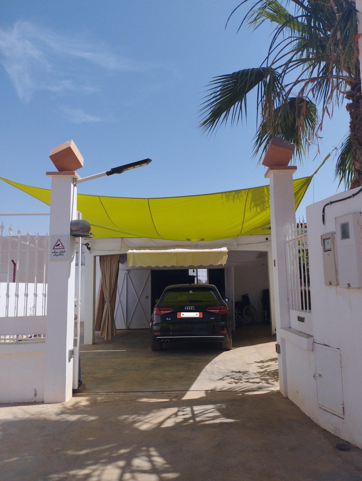 A covered carport is seen beneath a bright yellow canopy, providing shade for a vehicle. The entrance is framed by white walls and decorative columns, while a palm tree adds a touch of greenery to the scene.