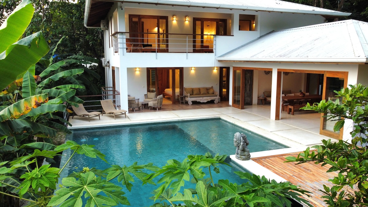 A modern villa is surrounded by lush greenery, showcasing a refreshing infinity pool. Lounge chairs are positioned beside the pool, providing a comfortable spot for relaxation. The large windows and outdoor seating area invite natural light and views of the tranquil outdoor environment.