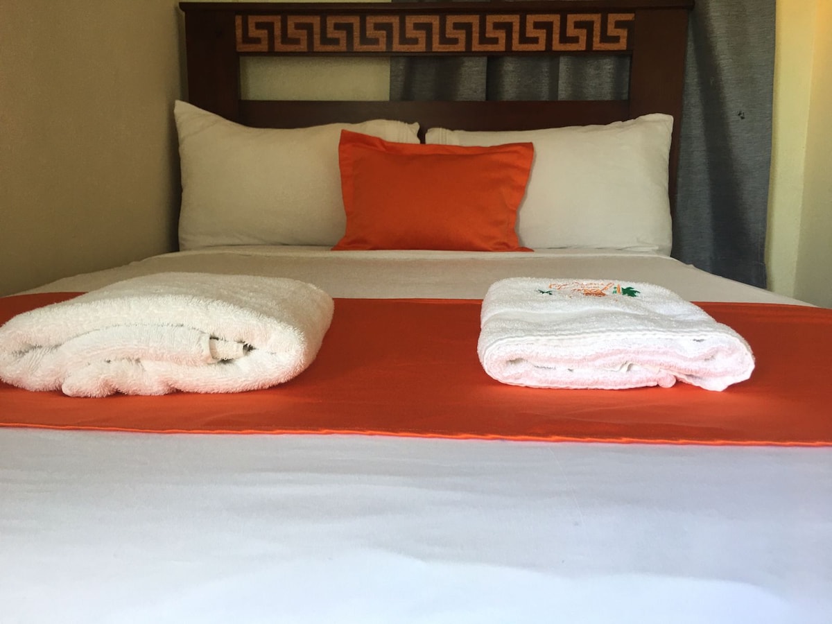 A comfortable bed is centered in the image, featuring two large white towels neatly rolled and positioned on a decorative orange bed runner. A bright orange pillow complements the arrangement against a backdrop of white linens and a patterned headboard.