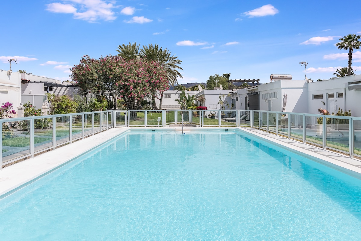 A clear swimming pool is surrounded by a glass barrier, reflecting the blue sky above. Lush palm trees and flowering plants are visible in the background, enhancing the serene landscape of the residential complex.
