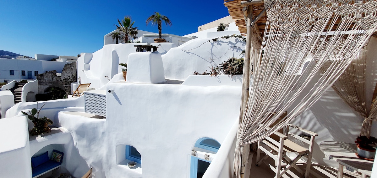 The image captures the whitewashed exterior of the traditional Santorinian architecture, showcasing curved surfaces and a blue sky above. Plants are observed on the terraces, and wooden furniture is visible, creating a serene outdoor space that invites relaxation.