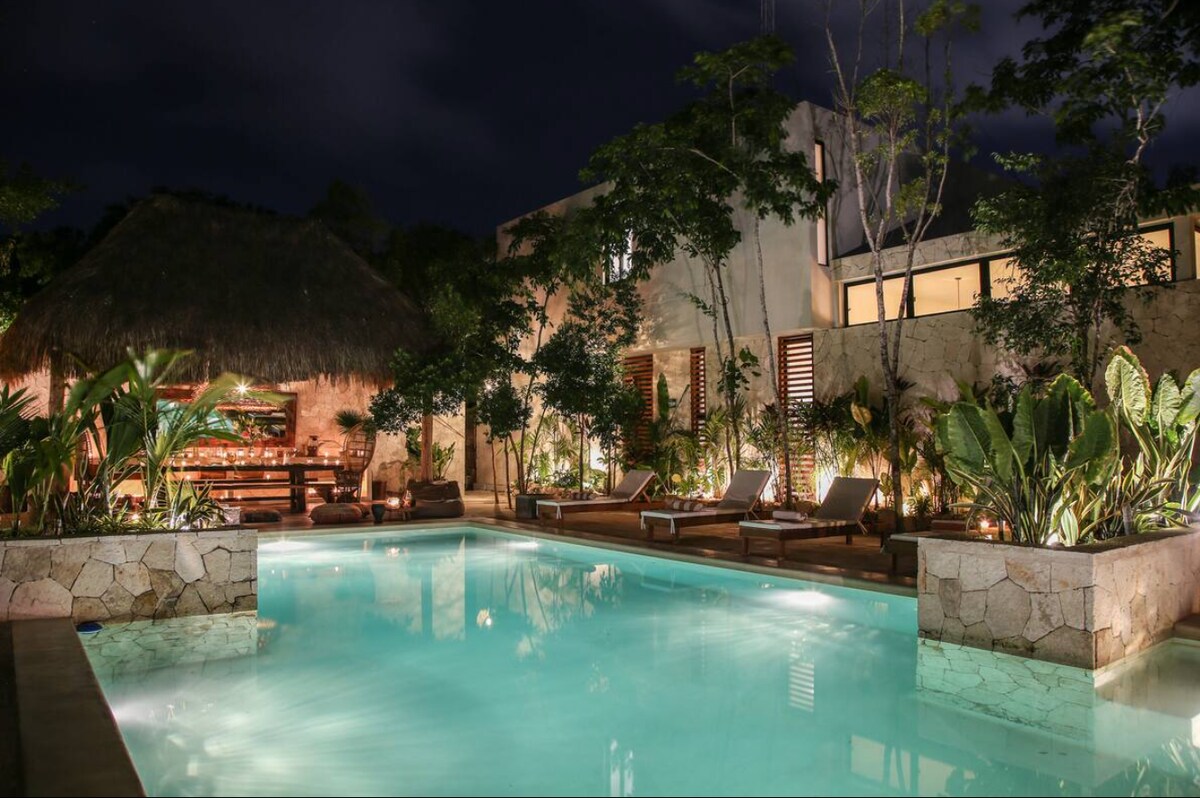 A serene pool area is illuminated at night, surrounded by lush tropical plants and stone walls. Comfortable lounge chairs are positioned along the water's edge, inviting relaxation. The nearby thatched-roof pavilion offers additional seating, enhancing the tranquil atmosphere amid the beautifully landscaped surroundings.