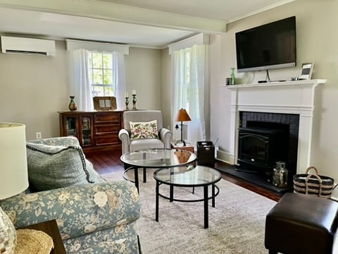 A comfortable living area is showcased with a floral-patterned couch and a glass coffee table. A television is mounted on the wall above a white fireplace. Natural light enters through the sheer curtains, illuminating the space that is complemented by a wooden cabinet and a small lamp.