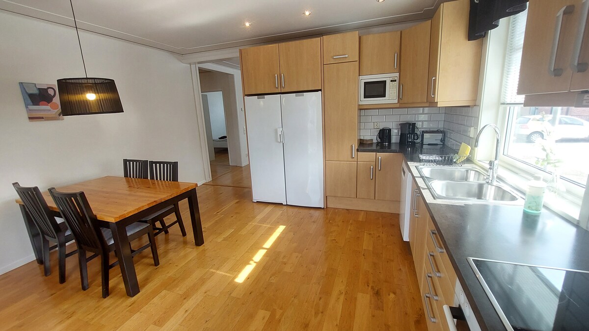 A modern kitchen area features wooden cabinetry and a stainless steel refrigerator. A dining table with four chairs is positioned to the side. Natural light fills the space, highlighting the light wood flooring and the organized countertop equipped with appliances like a microwave and coffee maker.