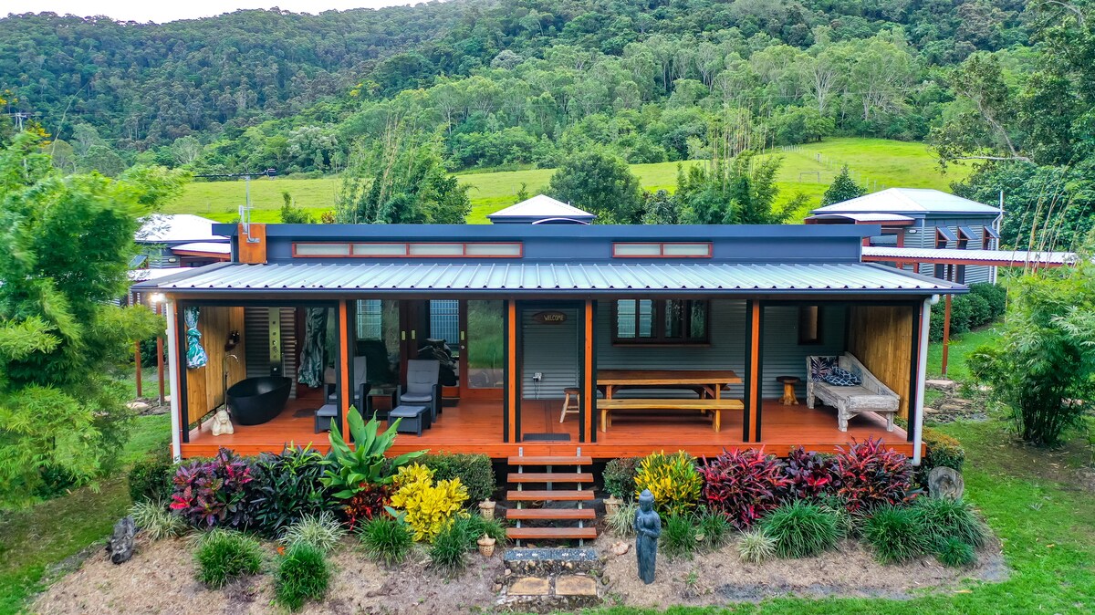 A wooden cabin is showcased among lush greenery, featuring a spacious veranda with comfortable seating. Vibrant plants decorate the front, and steps lead to the entrance. The backdrop consists of hills, creating a serene natural setting.