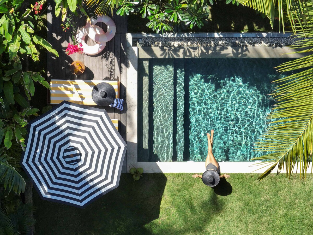 An aerial view captures a refreshing swimming pool surrounded by lush greenery. A striped sun umbrella is set near the pool, providing shade over lounge chairs. A guest is seen relaxing at the edge of the water, accentuating the tranquil outdoor setting.