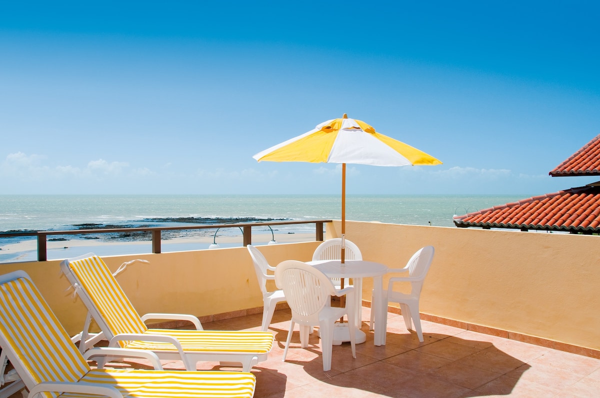 A private rooftop terrace is furnished with a table and four chairs under a large yellow umbrella. Two lounge chairs provide space for relaxation, while the expansive view overlooks the ocean and coastline, framed by a clear blue sky.
