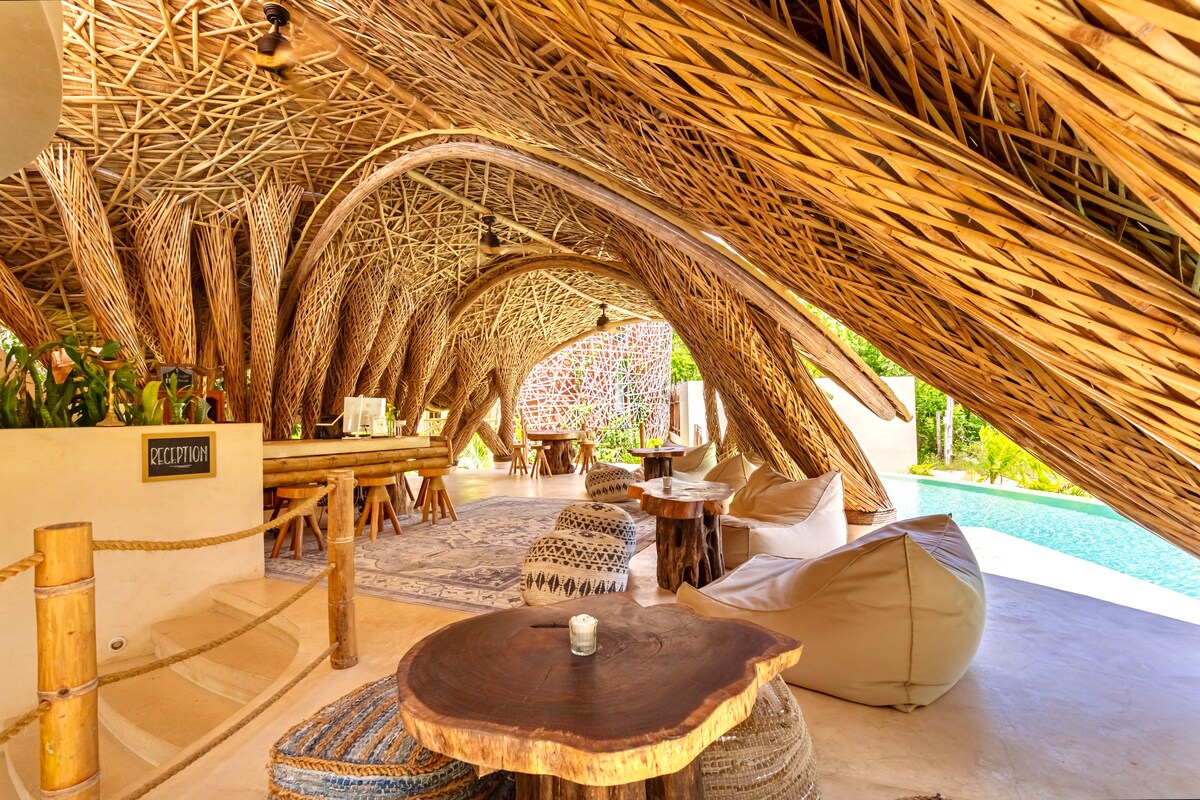 An open-air common area features a unique, organic structure made of woven bamboo. Comfortable seating arrangements include colorful poufs and wooden tables. Natural light floods the space, with lush greenery visible in the background, and a private pool adds a tranquil touch.