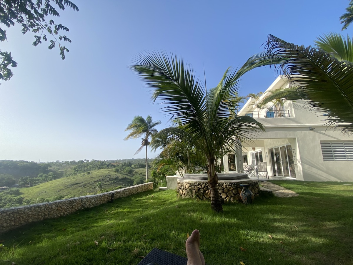 The exterior of a two-story villa is highlighted, surrounded by lush greenery and palm trees. A jacuzzi is visible on a stone patio, offering a view of the distant landscape and sky. The well-maintained lawn complements the tranquil setting.