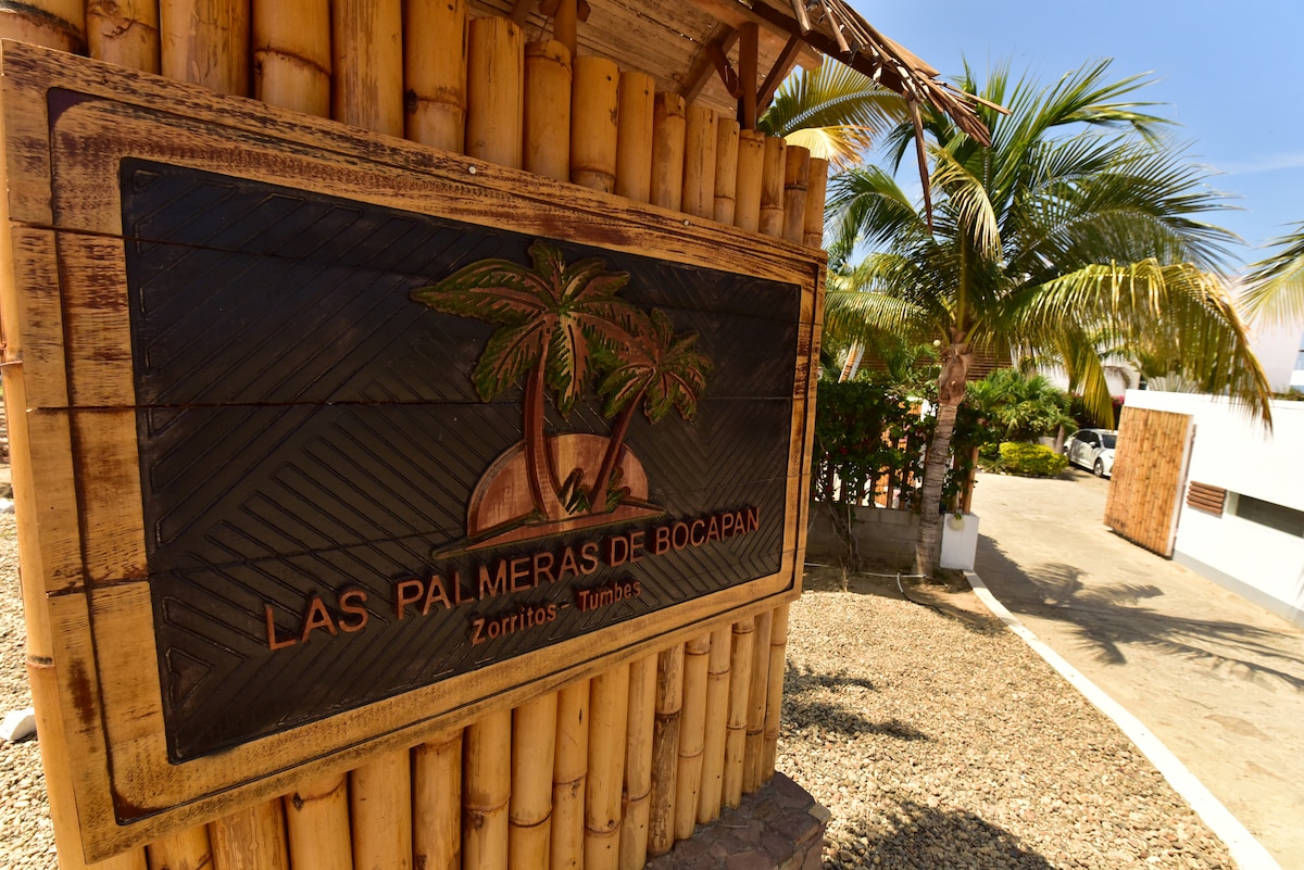 A wooden sign with the inscription 'Las Palmeras de Bocapan' is prominently featured. The sign is surrounded by tropical palm trees and gravel landscaping, showcasing the entrance to a welcoming beachside retreat.