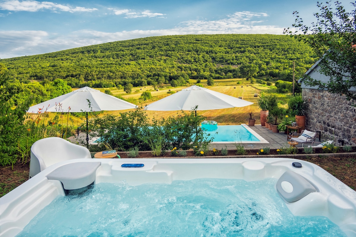 A hot tub is prominently featured in the foreground, with bubbling water inviting relaxation. In the background, a heated infinity pool is visible beneath shaded umbrellas, and lush green hills extend beyond, emphasizing the serene countryside setting.
