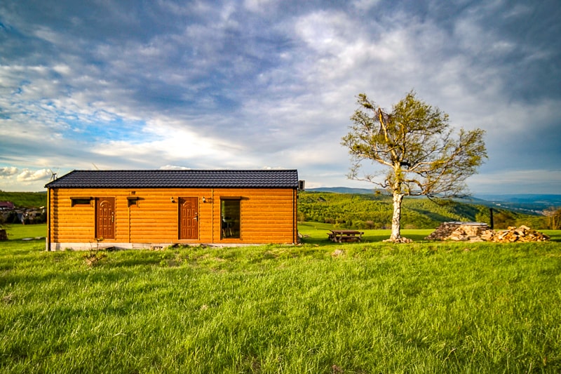 A wooden cabin is set on a grassy landscape, framed by a distant mountain view. Two doors are visible on the cabin's exterior, and a picnic table is positioned nearby. A single tree stands next to the cabin, adding to the natural surroundings.