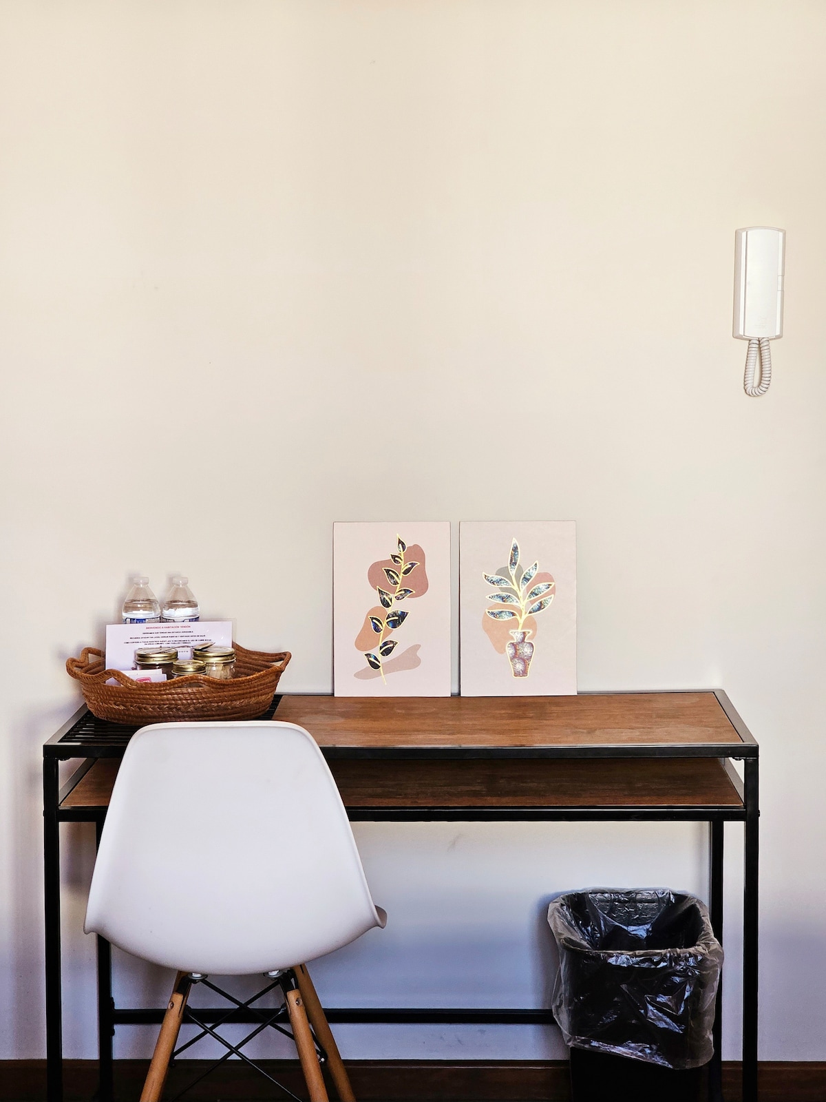 A small workspace is presented with a wooden desk and a white chair. Two art pieces featuring botanical designs adorn the wall above the desk. A basket filled with items sits on the desk, alongside two bottles of water, while a wastebasket is positioned near the floor.