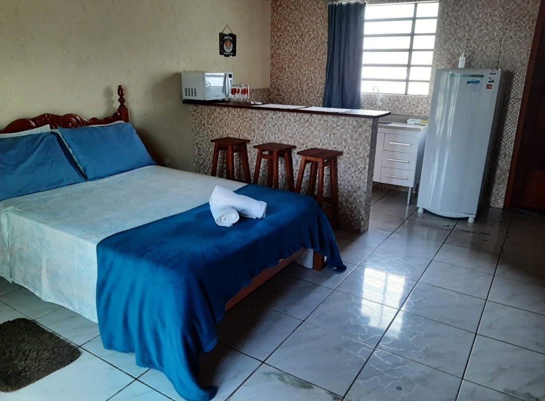 The loft's interior is illuminated by natural light, showcasing a spacious bed dressed in blue linens. A small seating area with wooden stools is positioned near the counter, with a refrigerator and microwave visible in the background, enhancing functionality and comfort.