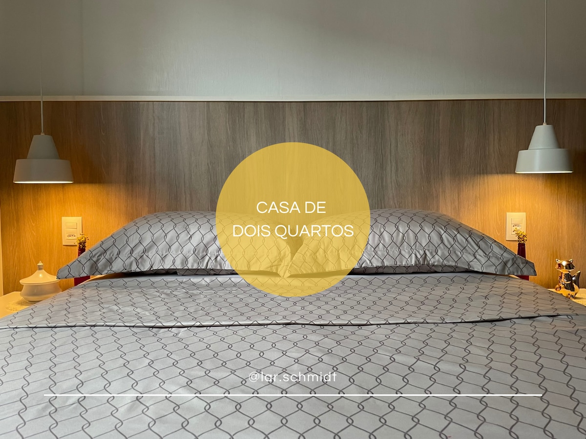 A spacious sleeping area features a neatly made bed with geometric-patterned linens. Soft lighting is provided by two pendant lamps on either side, creating a calming ambiance. The headboard is crafted from natural wood, complementing the serene color palette of the room.